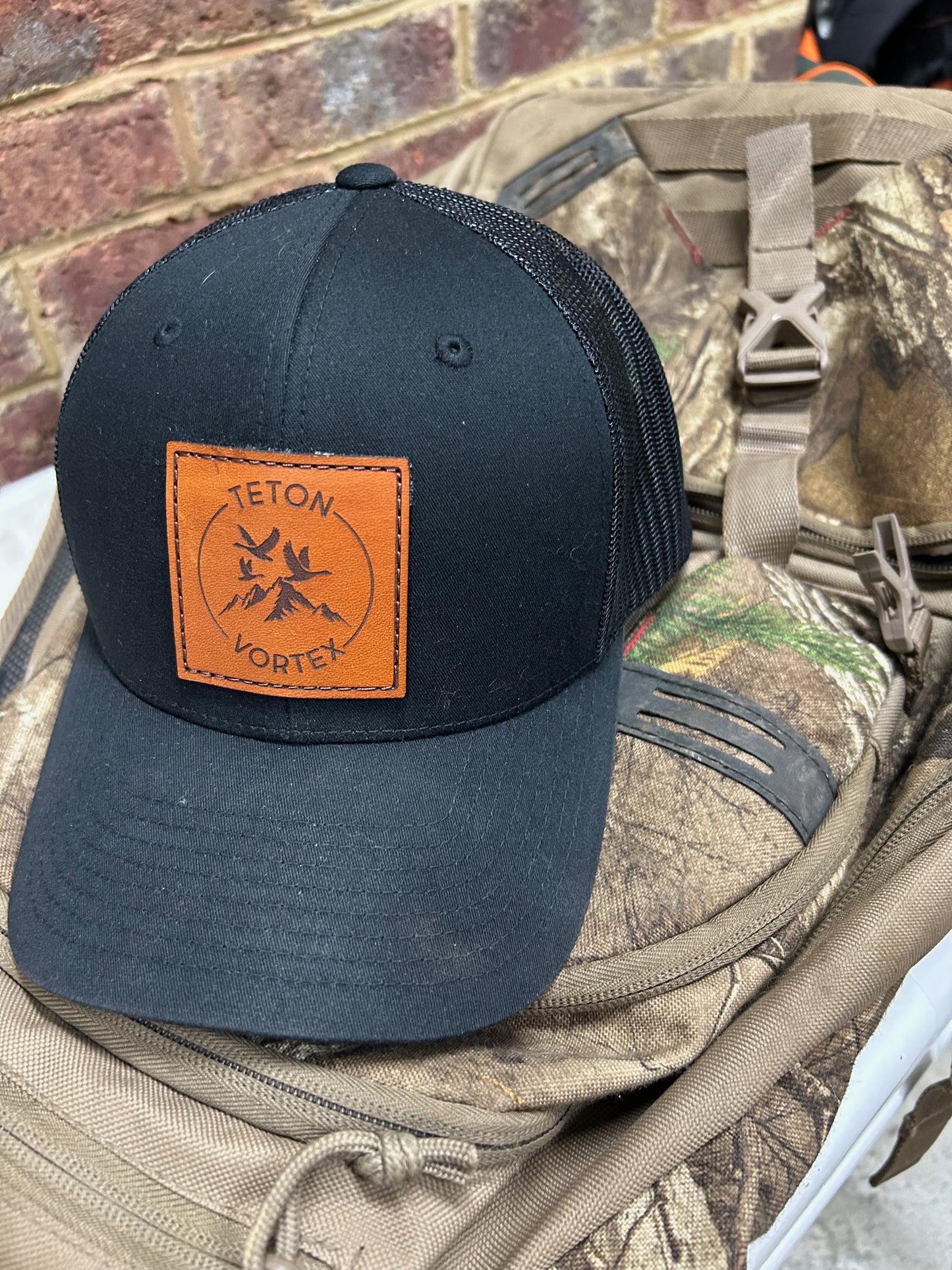 Black cap with brown leather Teton Vortex patch on a camouflage backpack against a brick wall.