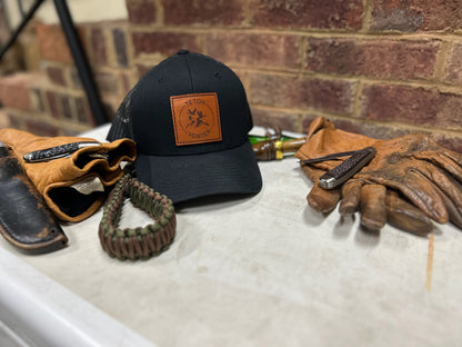 Black cap with Teton Vortex leather patch, gloves, and hunting knife on a concrete surface with a brick wall background.