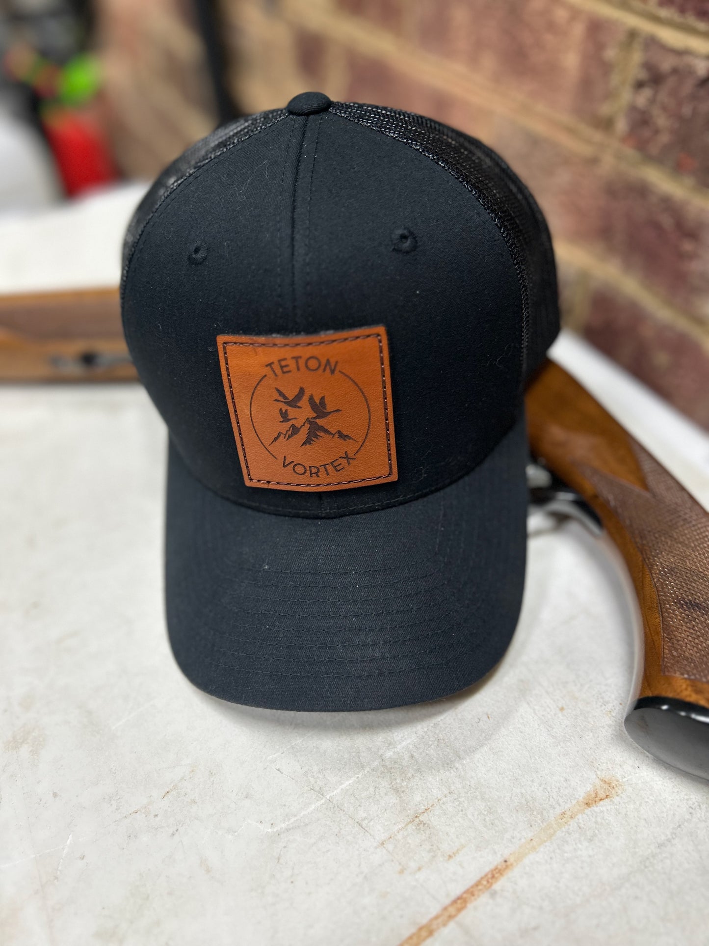 Black cap with a brown leather Teton Vortex patch next to hunting rifle on a white surface