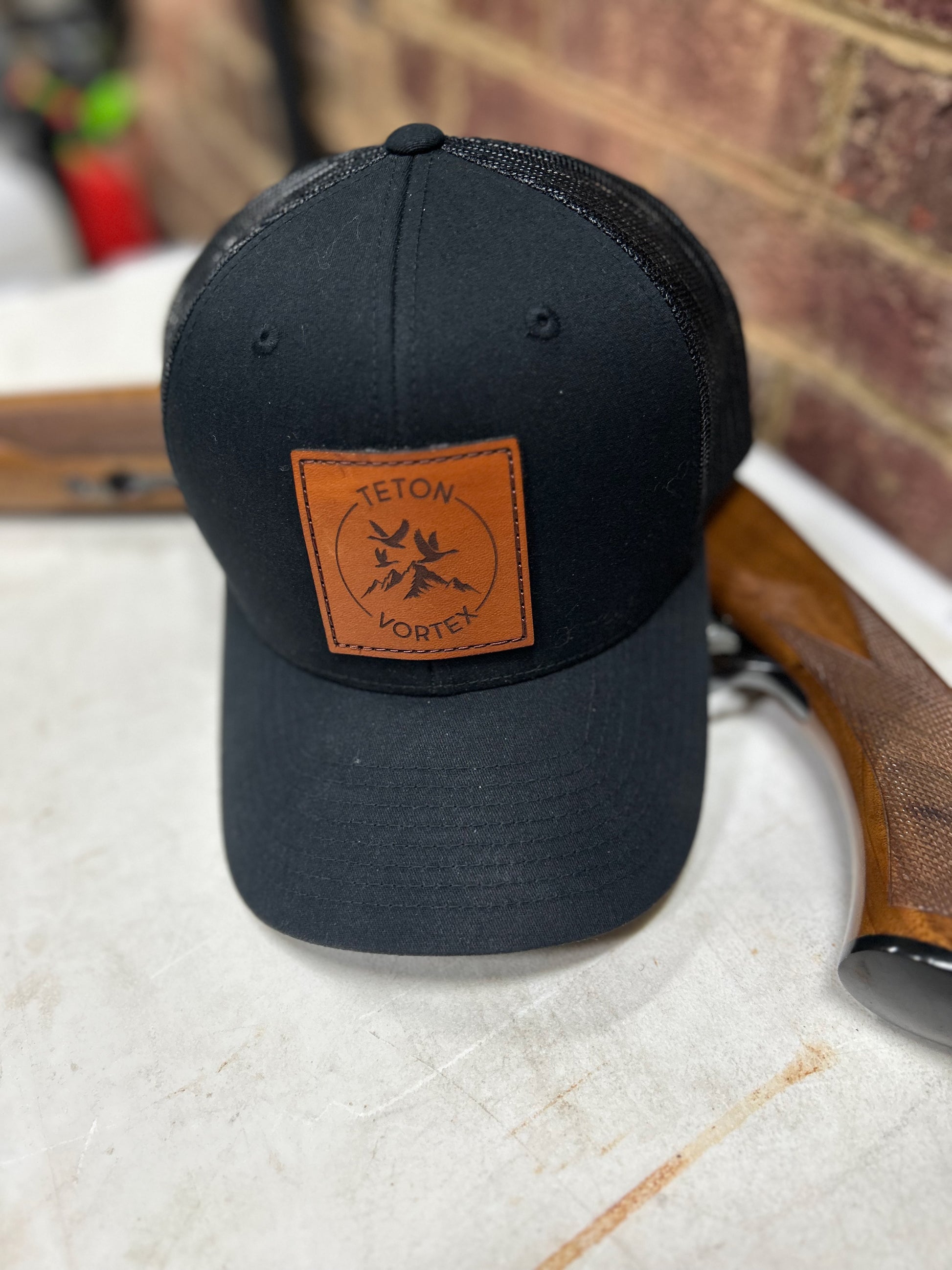 Black cap with a brown leather Teton Vortex patch next to hunting rifle on a white surface